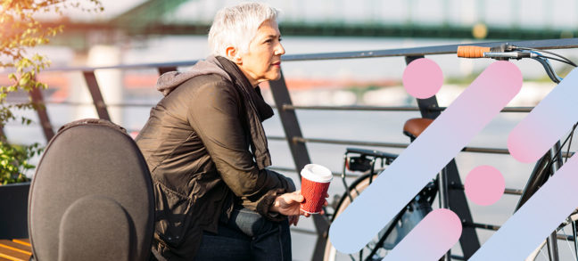 Older woman sits on bench overlooking a bridge and thinks deeply while holding coffee cup